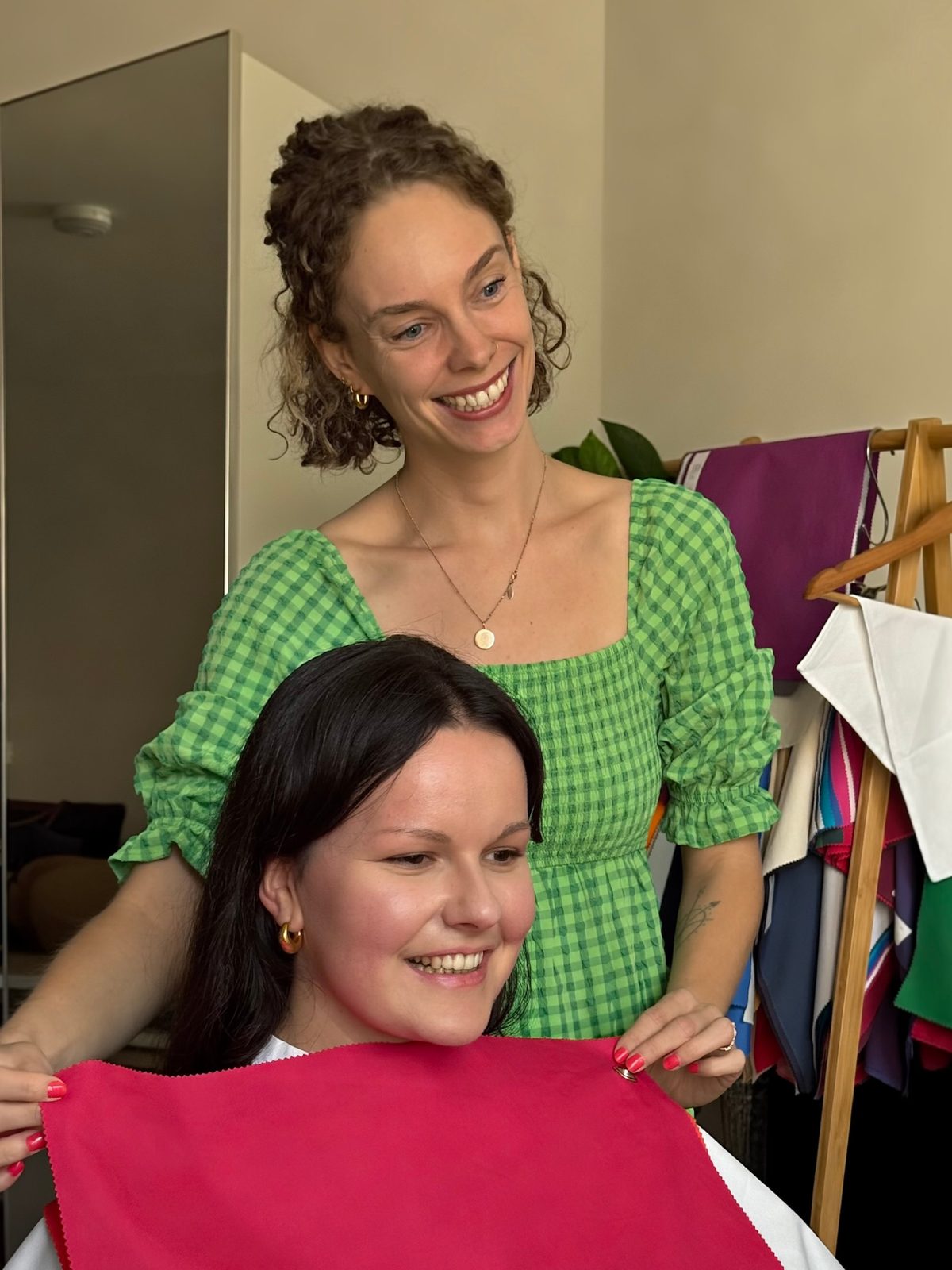 Shann holding a bright pink drape beneath a smiling client&rsquo;s chin as they both look towards the mirror